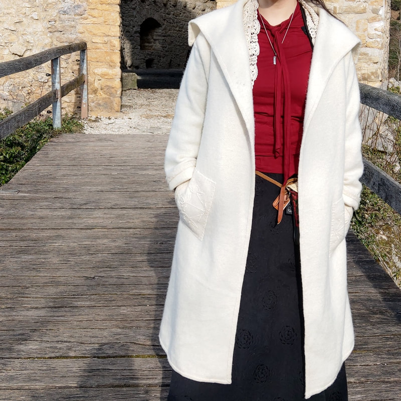 photo of Christina Serra modeling a Serra Designs long white wool jacket with white on white wool embroidered pockets, standing in front of pale yellow castle ruins.