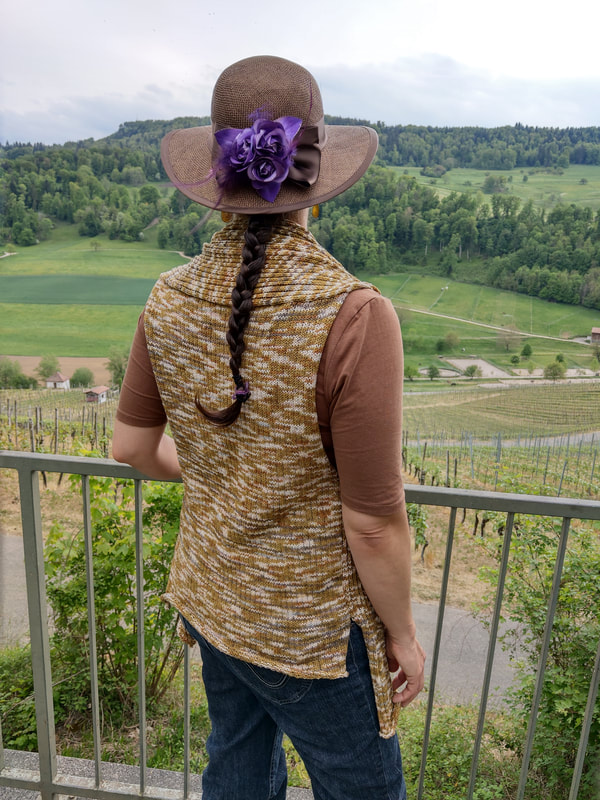 Photo of Christina Serra modeling Serra Designs brown sleeveless knit open cardigan sweater. She is facing away from teh camera, looking at a green hillside with horse pastures, vineyards and trees. 